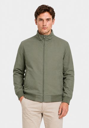 Mann steht nach vorne gerichtet, trägt eine zugezippt olivgrüne Jacke und beigefarbene Hose vor einem schlichten, hellen Hintergrund.