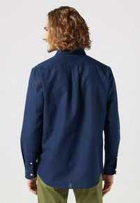 Camicia a maniche lunghe in cotone blu navy con colletto, polsini con un bottone, dettaglio con piega sulla schiena e orlo dritto. Pantaloni verdi visibili in basso.