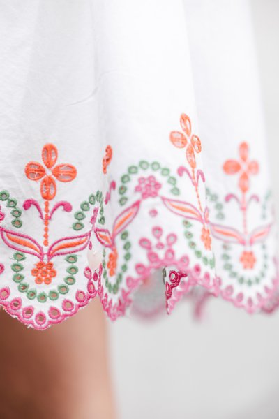 Λευκό ύφασμα με πολύχρωμη κεντητική σε πορτοκαλί, πράσινο και ροζ. Διαθέτει λουλουδένια σχέδι α και λεπτομέρεια με κυματιστή ραφή στο τελείωμα.