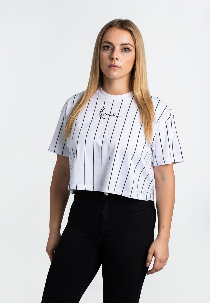 Gekürztes weißes T-Shirt mit schwarzen vertikalen Streifen, ausgestattet mit einem schwarzen Signaturlogo auf der Brust, kombiniert mit hoch-taillierten schwarzen Jeans.