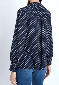 Blouse à motif marine avec un col arrondi, des manches longues bouffantes et des poignets boutonnés. Présente un motif géométrique subtil et un pli dans le dos.
