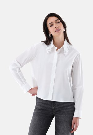 Frau trägt eine weiße Bluse mit langen Ärmeln und Spitzenkragen sowie dunkelgraue Jeans, posiert mit einer Hand an der Hüfte vor einfachem Hintergrund.