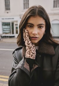 Bruine leren handschoenen met een luipaardprintpatroon, zachte textuur, gedragen door een persoon met donker haar, in een zwarte jas met een schapenwollen kraag.