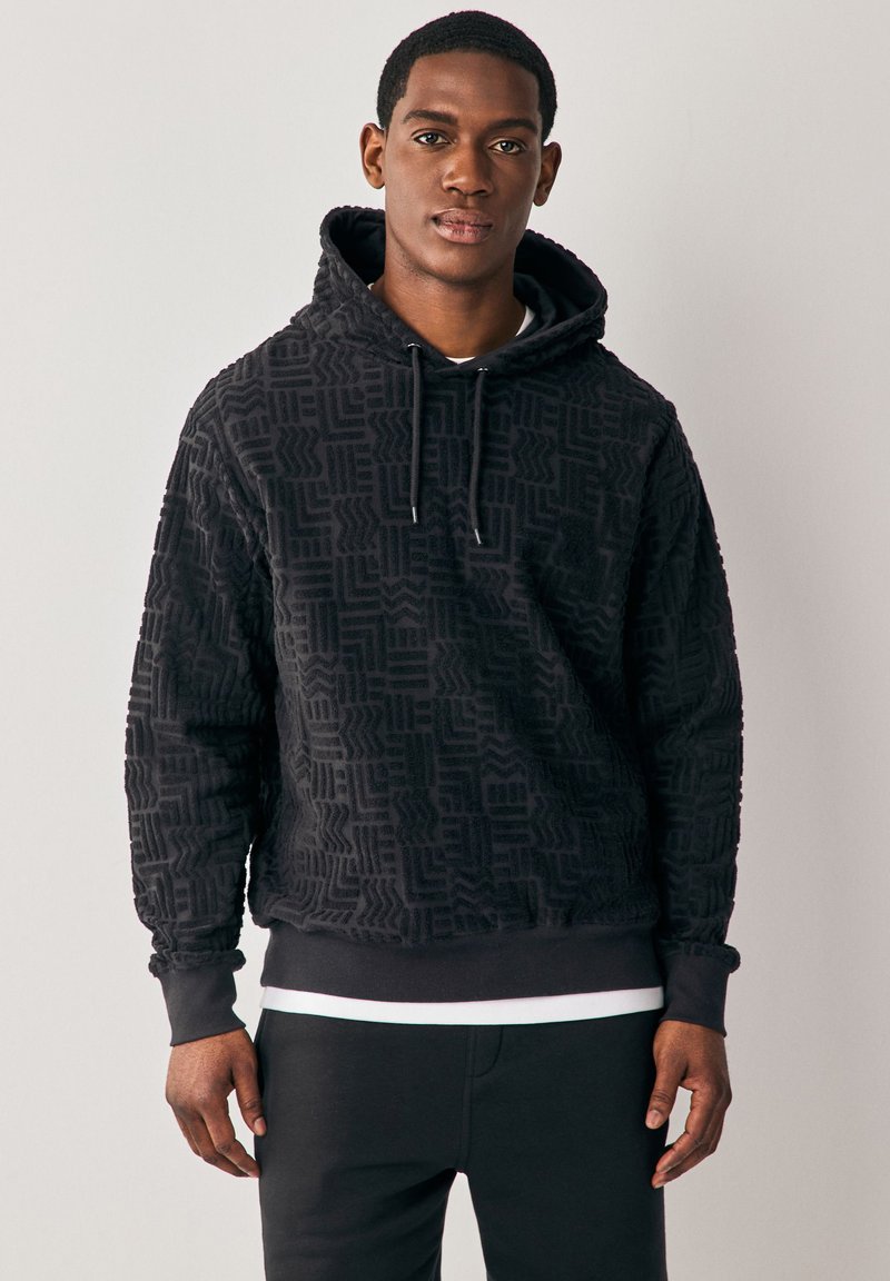 Svart hoodie med ett mönster i geometrisk struktur, med dragsko på huvan, ribbade muddar och en enfärgad svart midjeresår över smala byxor.