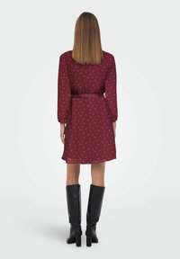 Femme aux cheveux raides mi-longs, portant une robe florale bordeaux et des bottes à talons noires jusqu'au genou, de dos sur un fond blanc uni.
