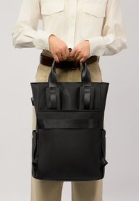 Czarna torba na ramię z nylonu z podwójnymi uchwytami u góry, dwiema bocznymi kieszeniami i płaskim frontem. Gładka tekstura i minimalistyczny design.