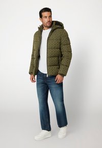 Green patterned puffer jacket with a hood, zip closure, ribbed cuffs, paired with blue jeans and white sneakers, standing against a light background.