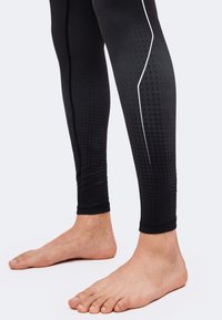 Des pieds nus portant des leggings noirs avec un motif discret à pois et un détail de ligne blanche courbe sur le bas des jambes sur un fond clair.