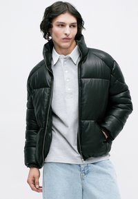 Jachetă puffer din piele neagră, cu guler înalt, închidere cu fermoar și mansete elastice, purtată peste un tricou polo gri deschis și blugi albaștri deschiși.
