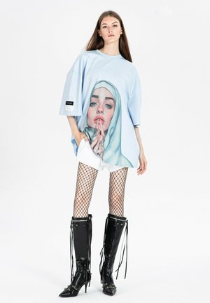 Joven con camiseta azul claro de gran tamaño con un gran gráfico de una mujer rezando con pañuelo en la cabeza, medias de rejilla, pantalones cortos blancos y botas negras hasta la rodilla.