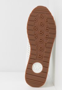 Semelle d'une chaussure blanche avec une semelle extérieure marron arborant des logos "MK" répétés et un emblème circulaire "MK" près du talon.