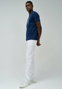 Camiseta de algodón azul marino con gráfico blanco, combinada con pantalones vaqueros de algodón blancos y zapatillas blancas. El modelo posa con las manos detrás de la espalda, mirando ligeramente de lado.