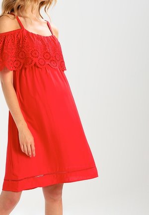 Kobieta w czerwonej sukience bez rękawów z koronkową nakładką w kwiatowy wzór i ramiączkami, stojąca na tle jednolitego jasnego tła.