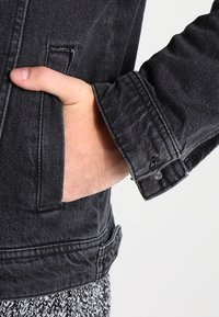 Svart denimbomulljacka med långa ärmar, med en knapp på manschetten, sydda fickor och ett strukturerat tyg. Handen placerad i fickan.