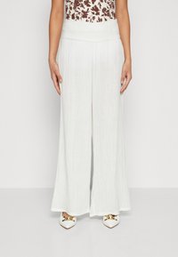Bílé kalhoty širokého střihu z texturované látky, s gumovým pasem a volným designem. Ve spojení s bílými špičatými botami se zlatými akcenty.