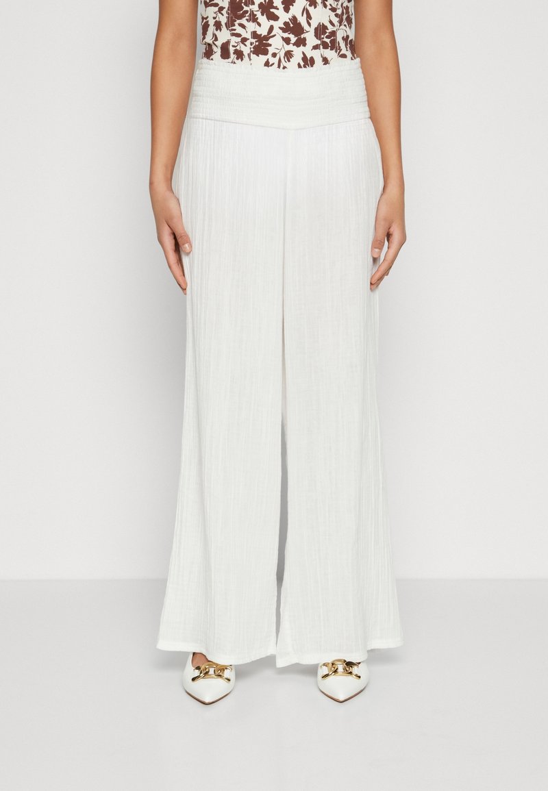 Bílé kalhoty širokého střihu z texturované látky, s gumovým pasem a volným designem. Ve spojení s bílými špičatými botami se zlatými akcenty.