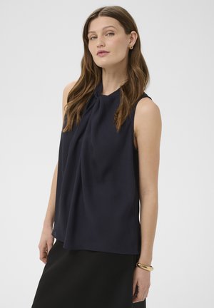 Vrouw met lang bruin haar, gekleed in een mouwloze marineblauwe blouse en een zwarte rok, geaccentueerd met gouden oorbellen en een armband.