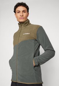 Fleecejakke med lynlås foran, høj krave og to sidelommer; farveblokdesign i grøn og brun med logo.