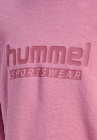 Różowy materiał z wypukłym logo „hummel” i napisem „SPORTSWEAR” w tym samym odcieniu różu na bluzie.