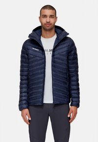 Granatowa pikowana kurtka puchowa z kapturem, z przeszytym wzorem, zapinanymi na zamek kieszeniami bocznymi oraz logo Mammut na piersi i rękawie.