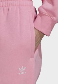 Rosa Adidas-träningsbyxor med elastisk midjeband, sidfickor och vitt broderat logotyp på vänster ben. Mjuk tygstruktur.