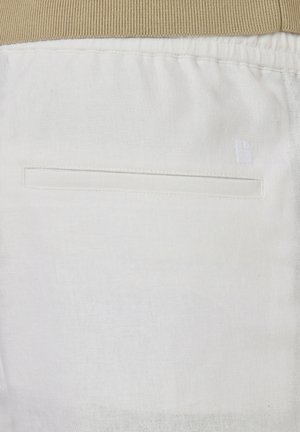 Gros plan sur un pantalon en tissu blanc avec une poche fendue sur le côté et une ceinture élastique beige visible en haut.