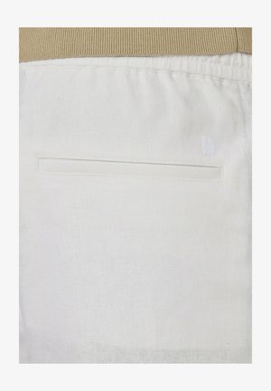 Gros plan sur un pantalon en tissu blanc avec une poche fendue sur le côté et une ceinture élastique beige visible en haut.