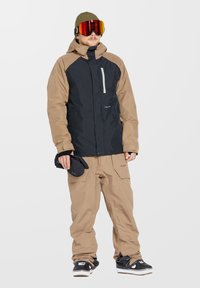 Snowboardoutfit met een zwart en bruin waterdichte jas, bruine cargobroek en zwarte schoenen. Accessoires zijn onder andere een bril met rode tint en een muts.