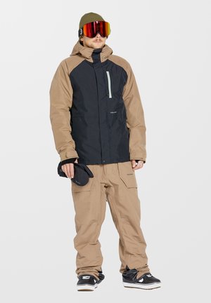 Conjunto de snowboard que incluye una chaqueta impermeable negra y marrón, pantalones cargo marrones y botas negras. Los accesorios son unas gafas de esquí con tinte rojo y un gorro.