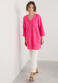 Roze linnen tuniek met een V-hals en knoopdetail aan de voorkant, gecombineerd met witte broeken en bruine sandaaltjes met bandjes. Eenvoudige, ontspannen pasvorm.