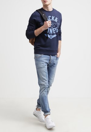 Młody mężczyzna w granatowej bluzie z grafiką, jasnoniebieskich dżinsach, białych trampkach, noszący na jednym ramieniu czarny plecak.