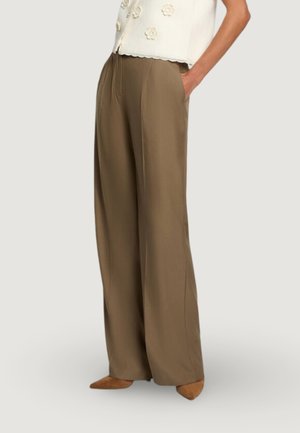 Žena oblečená v hnědých širokých kalhotách s vysokým pasem, bílém bezrukávovém topu se zvlněným okrajem a hnědých špičatých botách, s jednou rukou v kapse.