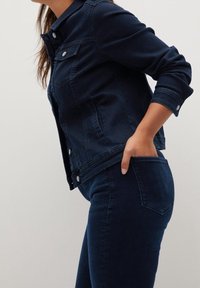 Mujer con chaqueta de mezclilla azul oscuro y vaqueros, con la mano en el bolsillo trasero, vista lateral sobre un fondo liso.