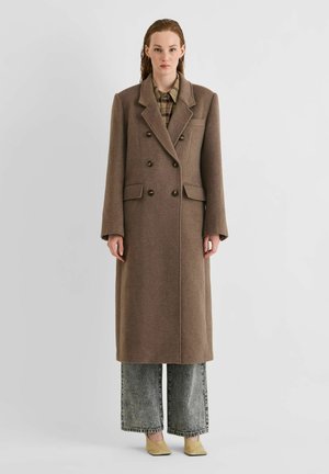 Jeune femme portant un long manteau marron à double boutonnage, une chemise à carreaux, un jean large gris et des chaussures beiges, debout devant un fond uni.