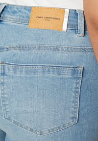 Jean en denim bleu clair avec une poche arrière carrée, un patch en cuir marron et des éléments en métal argenté sur la taille.