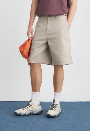 Smėlio spalvos krovinių šortai su priekinėmis kišenėmis, dėvimi su bordo marškinėliais ir baltais kojinaitėmis. Asmuo dešine ranka laiko oranžinę kepurę.