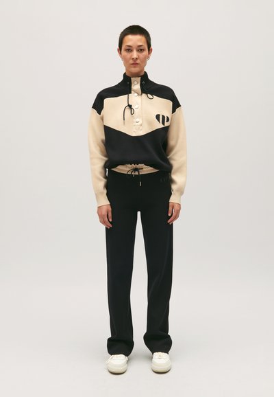 Pull en polaire noir et beige avec un col haut, une patte de boutonnage et un logo. Associé avec un pantalon de survêtement noir et des chaussures blanches.