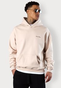 Helle rosa Kapuzenjacke aus weichem Material, mit einer Vordertasche, gerippten Bündchen und dezentem Branding auf der Brust. Getragen mit schwarzen Hosen.