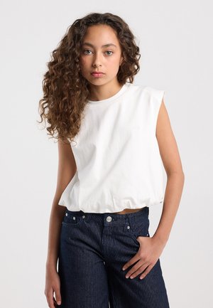 Jeune femme aux longs cheveux bouclés portant un haut blanc sans manches et un pantalon rayé sombre taille haute, debout avec la main dans la poche.