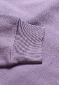 Violetinis megztinis su raižyta rankove ir lygiu, minkštu paviršiumi. Audinys atrodo tamprus ir lengvas, pabrėždamas savo konstrukciją.