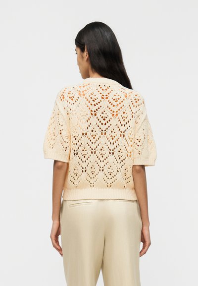 Femme aux longs cheveux foncés portant un pull tricoté crème à manches courtes avec des motifs en losange et un pantalon beige clair, vue de dos.