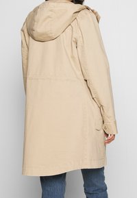 Beżowa, długa kurtka z kapturem, zapinana na guziki z bocznymi kieszeniami i luźnym krojem. Wykonana z lekkiego materiału o gładkiej teksturze.