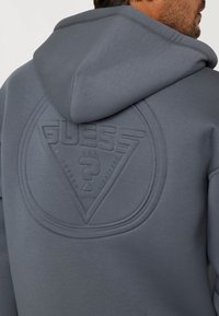 Sudadera con capucha gris que presenta un parche de logo en relieve con un diseño triangular en la parte posterior, con una textura suave y detalles de costura.