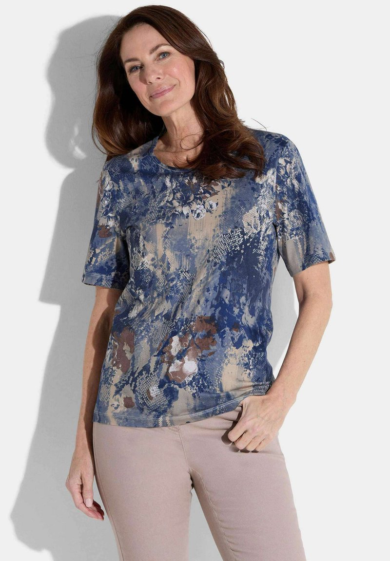 Femme aux longs cheveux bruns portant un haut à manches courtes bleu et beige à motifs et un pantalon rose clair, se tenant avec une main sur la hanche.