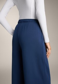 Temno modri široki hlače z elastičnim pasom, narejene iz mehkega materiala. Nad hlačami je oblečena dolga belinga majica.
