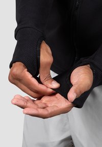 Schwarze Sportjacke mit gerippten Bündchen, die Daumenlöcher aufweist. Die Hände greifen die Bündchen, während die Jacke unzipped ist.