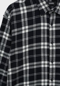 Camicia a quadri bianca e nera con bottoni e colletto, che mostra la trama del tessuto e l'etichetta del marchio Pull&Bear all'interno del colletto.