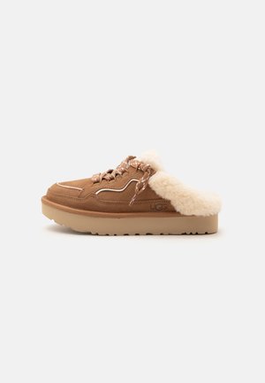 Bruine suède instapschoen met dikke zool, crème bontvoering, gemêleerde veters en een geperst UGG-logo aan de zijkant, zijaanzicht.