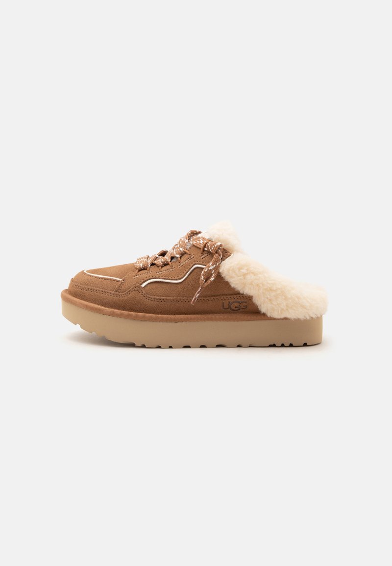 Καφέ δερμάτινο παπούτσι slip-on με παχιά σόλα, επένδυση από κρεμ γούνα, ραμμένα κορδόνια με σχέδιο και ανάγλυφο λογότυπο UGG στο πλάι, εμφανίζεται στο πλάι.