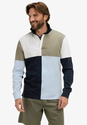 Homme souriant, portant un pull à manches longues avec une fermeture éclair jusqu'au quart, à blocs de couleurs blanc, beige, bleu marine et bleu clair, associé à un short vert olive.